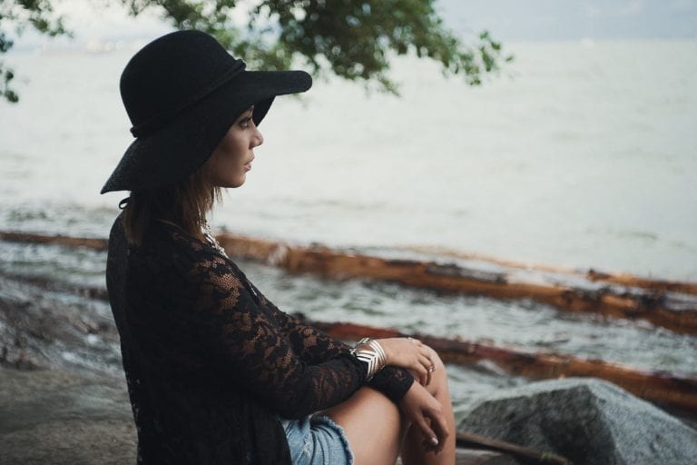 Natural light portrait of a young woman in a black hat sitting on a rock under a tree looking out over English Bay