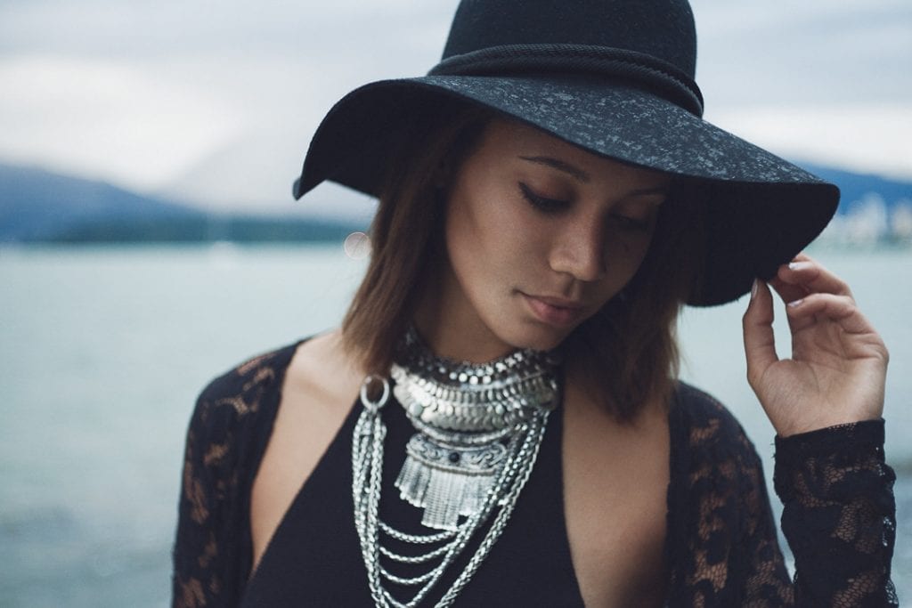 Natural light portrait of a young woman in a black hat and wearing stacked silver necklaces looking down with English Bay in the background