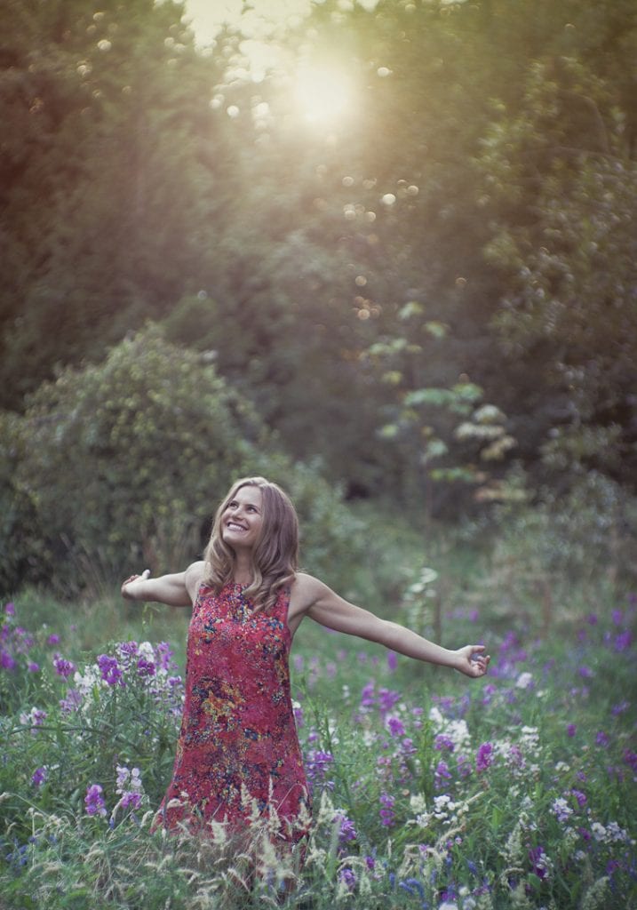 Natural light image of a young woman with outstretched arms in a field of wildflowers backlit by the setting sun by Vancouver contemporary portrait photographer Angela McConnell