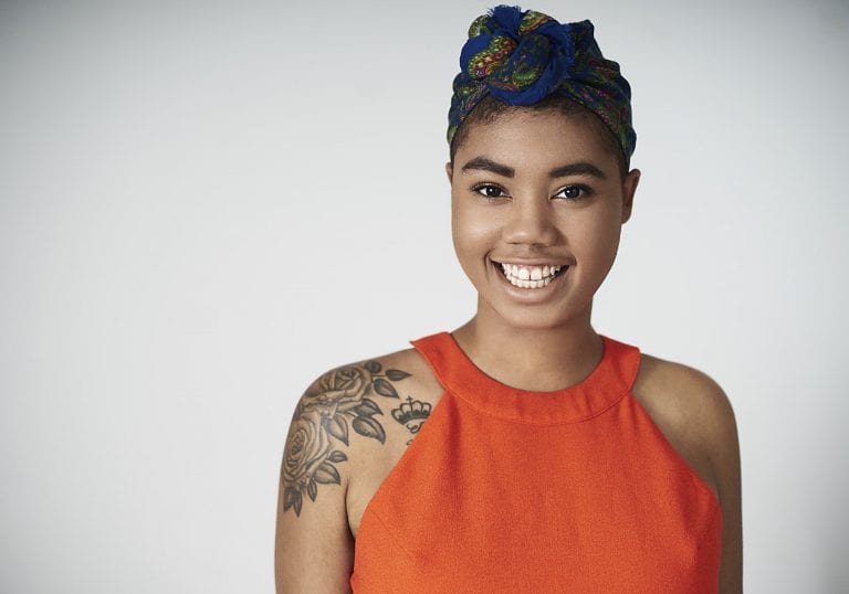 Studio light portrait of a young black woman with tattoos wearing a bright orange dress with a blue patterned head scarf smiling at the camera by Vancouver contemporary portrait photographer Angela McConnell