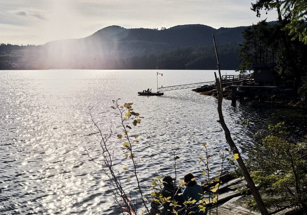 Image of a dock with deck chairs and a Canadian flag on an inlet with the sunshine streaming over the hills in Autumn at Sechelt on the Sunshine Coast, BC