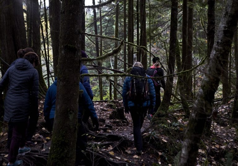 Women with backpacks walking through a forest path surrounded by trees covered in hanging moss during a self care and fitness workshop by Vancouver workshop and retreat photographer Angela McConnell