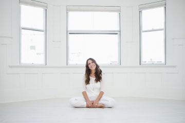 A yoga teacher sits in easy pose in a white room in front of windows at True Nosh Yoga by Vancouver business portrait and branding photographer Angela McConnell