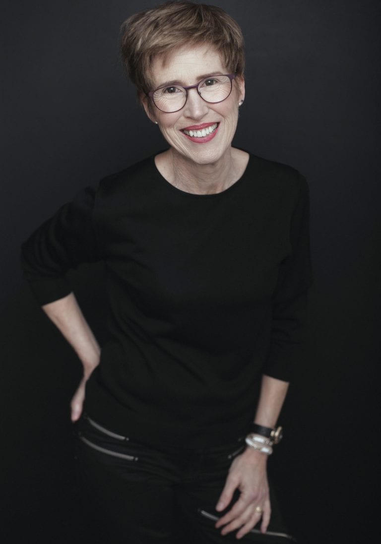 Older woman dressed in black and wearing glasses smiles during a portrait session by Vancouver contemporary portrait photographer Angela McConnell